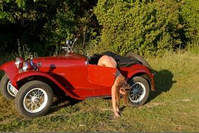 White solo girl poses naked in and on a vintage roadster out among the trees